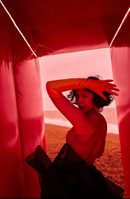 Portrait of a woman in a red inflatable structure on a beach, wearing orange gloves, with an ocean view.