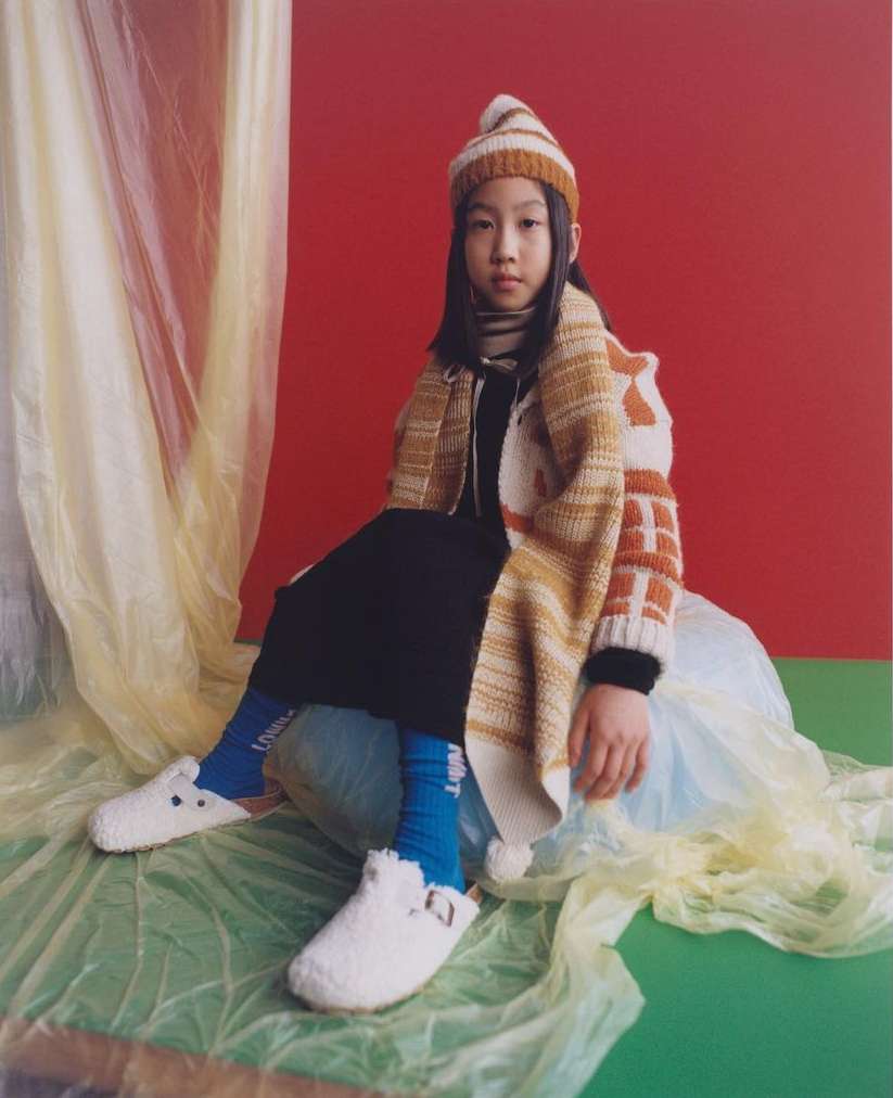Young Asian girl in striped knit hat and cardigan sits in studio on plastic wrap, wearing blue socks and white furry clogs.