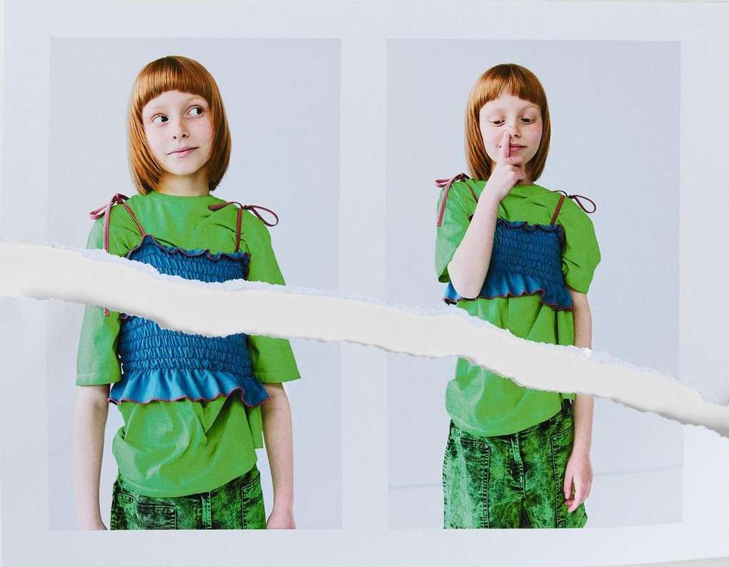 Two photos of a child model with red hair wearing a green t-shirt and blue top with green denim shorts against a white background.
