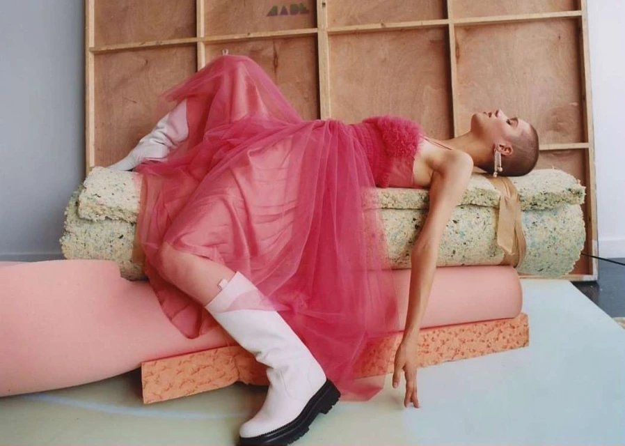 Woman in pink dress and white boots poses on foam blocks in a photo studio with wood-paneled background for a fashion editorial.