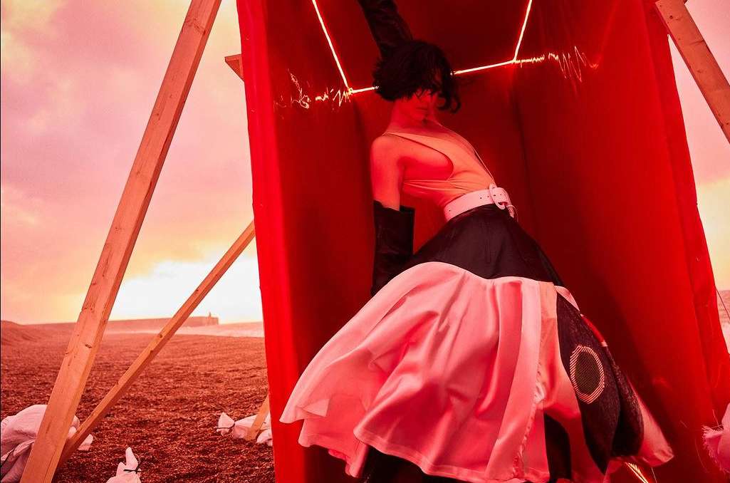Fashion editorial shot of a woman in an avant-garde dress inside a red box on a beach with a cloudy sky at sunset.
