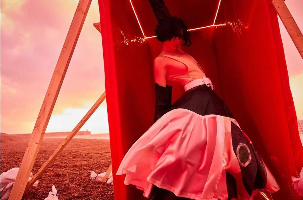 Fashion editorial shot of a woman in an avant-garde dress inside a red box on a beach with a cloudy sky at sunset.