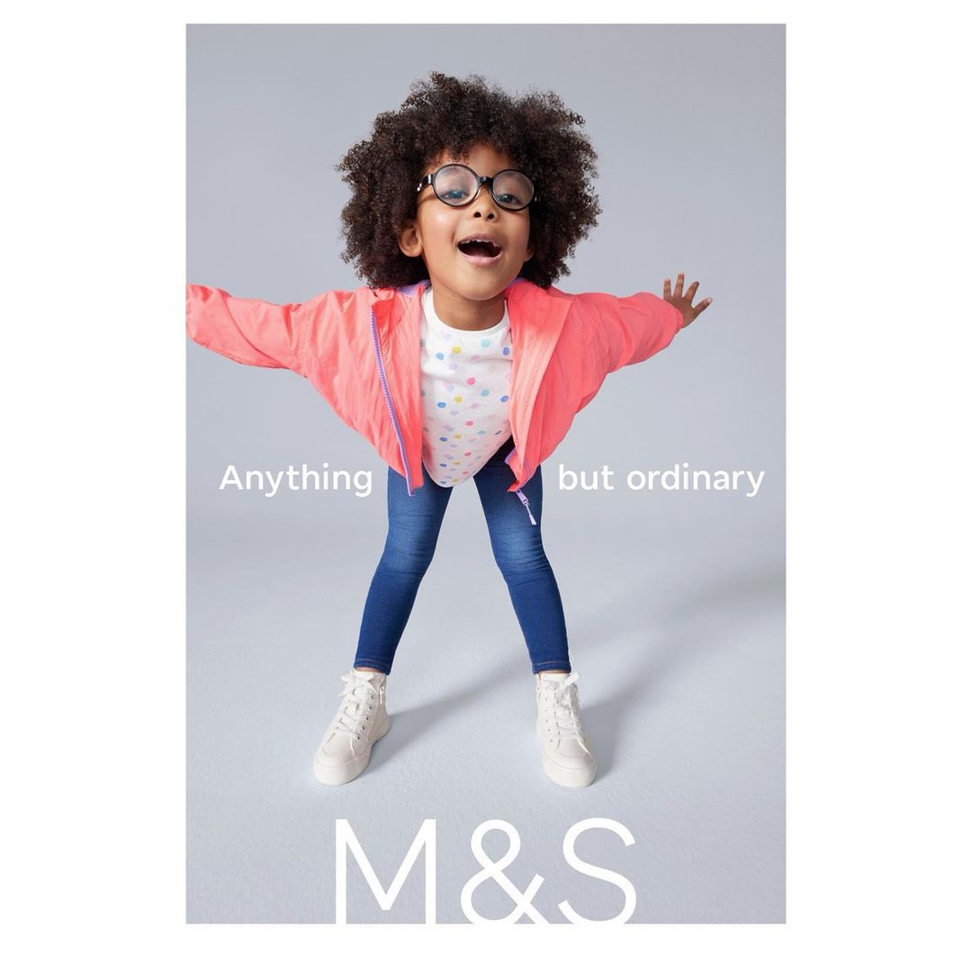 Adorable child model in a playful pose, wearing round glasses, a bright pink jacket, and jeans, set against a simple grey background.