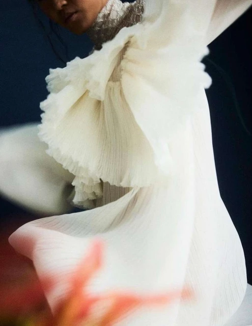 Close-up fashion shot of a person wearing a light and flowing ruffled gown against a dark background, emphasizing texture and movement.