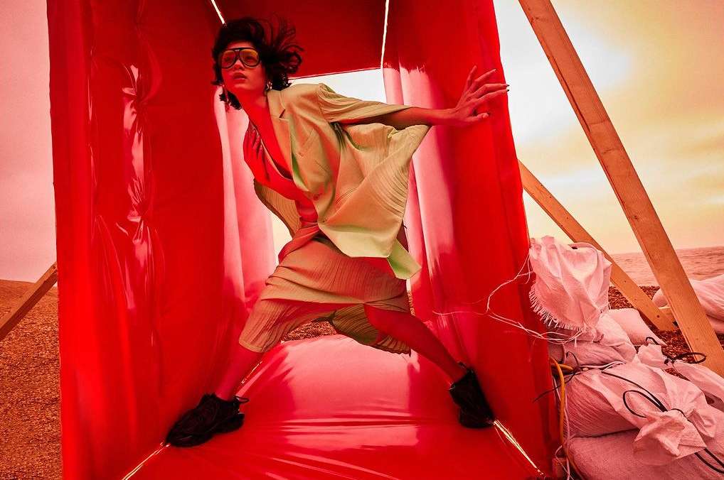 Fashionable person in green suit and sunglasses stands in a red vinyl tunnel structure on a beach with sand bags visible.