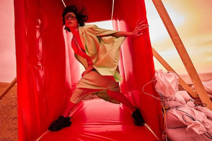 Fashionable person in green suit and sunglasses stands in a red vinyl tunnel structure on a beach with sand bags visible.