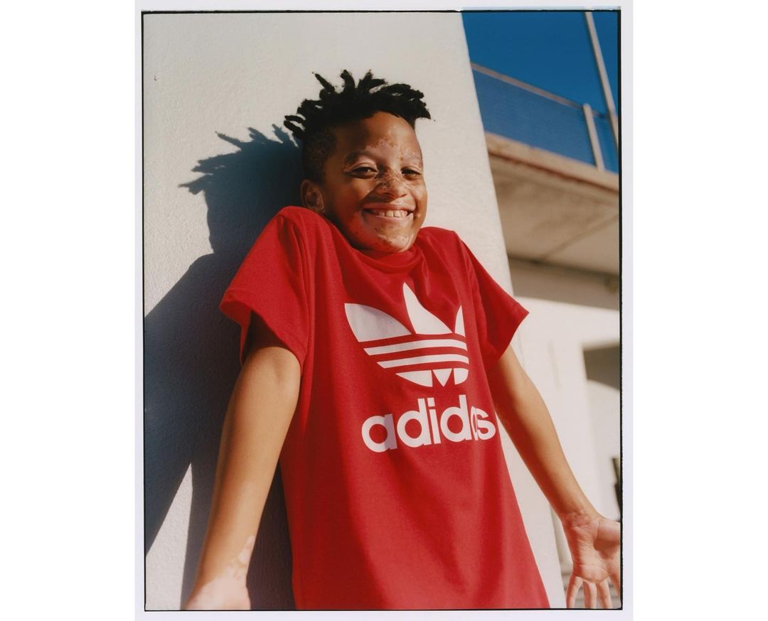 Smiling boy in a red Adidas t-shirt, standing outdoors next to a white wall under a blue sky. He has short dark hair.