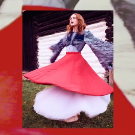 A woman with red hair wearing a vibrant red skirt spinning outdoors in front of a rustic wooden building.