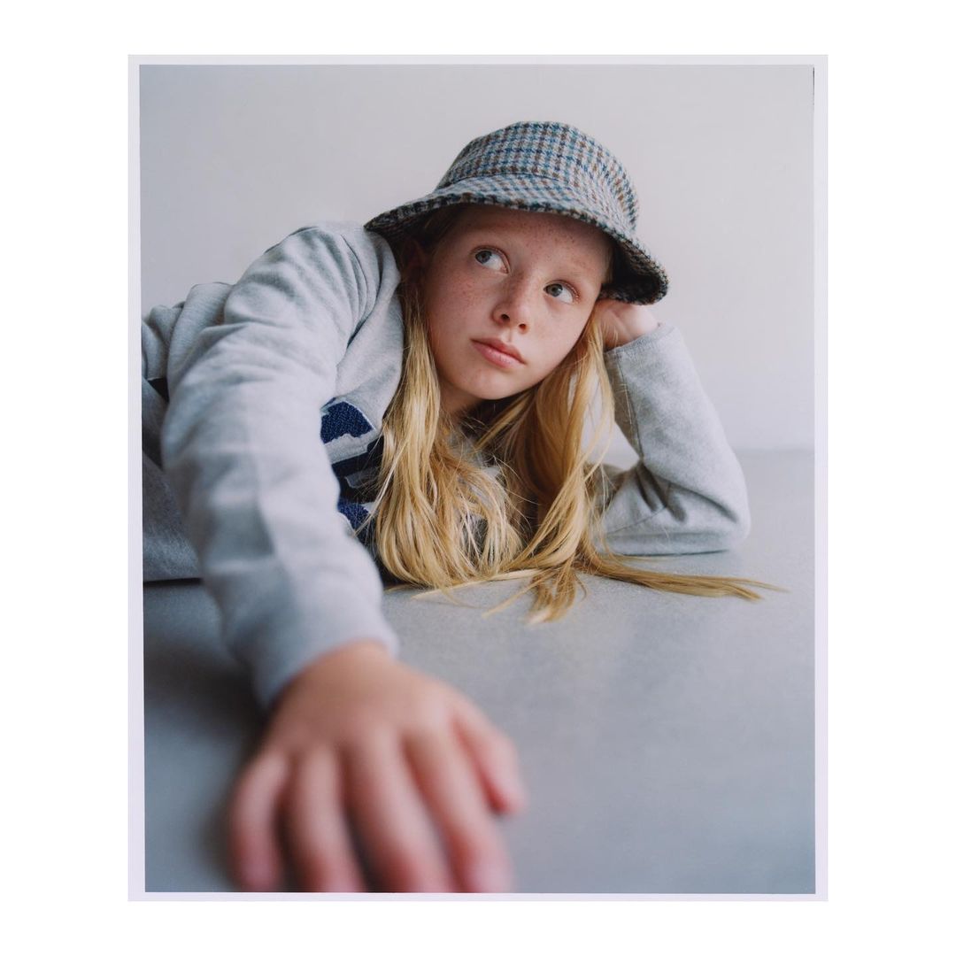 Young girl with blonde hair wearing a grey sweatshirt and patterned bucket hat lying on a grey table, looking away from the camera.