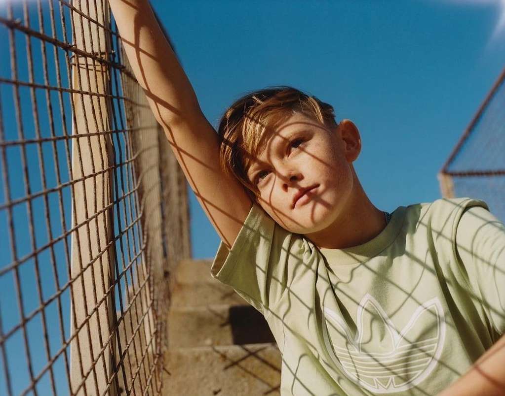 Low angle view of a young boy with blond hair wearing a t-shirt with the Adidas logo, leaning against a wire fence under a blue sky.