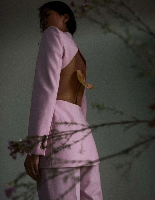 Back view of an adult female posing in a pink jacket and pants, open back with a leaf, soft lighting against a neutral background.
