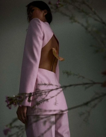 Back view of an adult female posing in a pink jacket and pants, open back with a leaf, soft lighting against a neutral background.