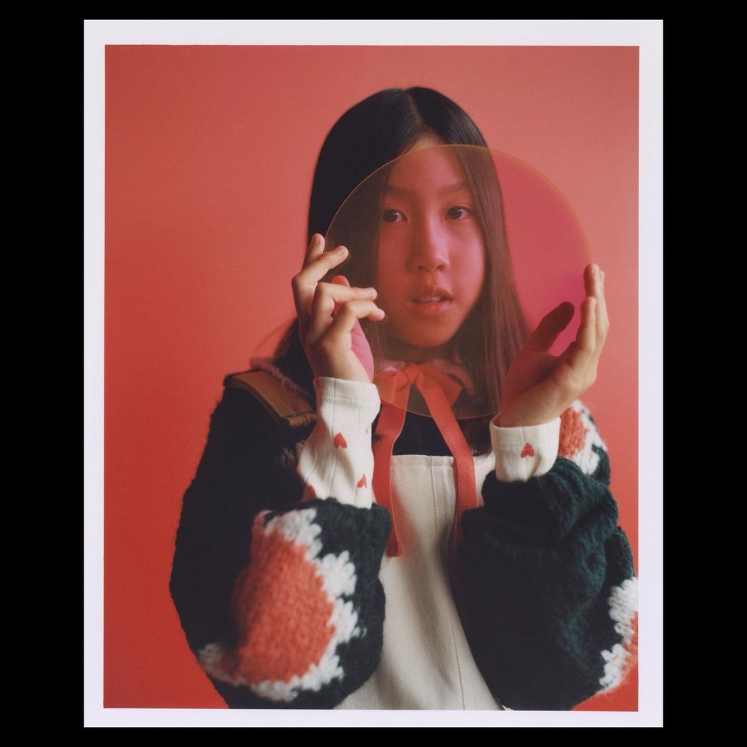 Young Asian woman poses indoors with clear pink discs in front of a red background, wearing a black and white patterned sweater.
