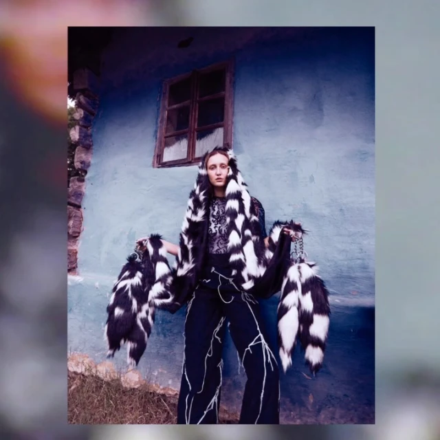 A person wearing a black and white fur hooded shawl and pants standing outdoors in front of a light blue house with a window.