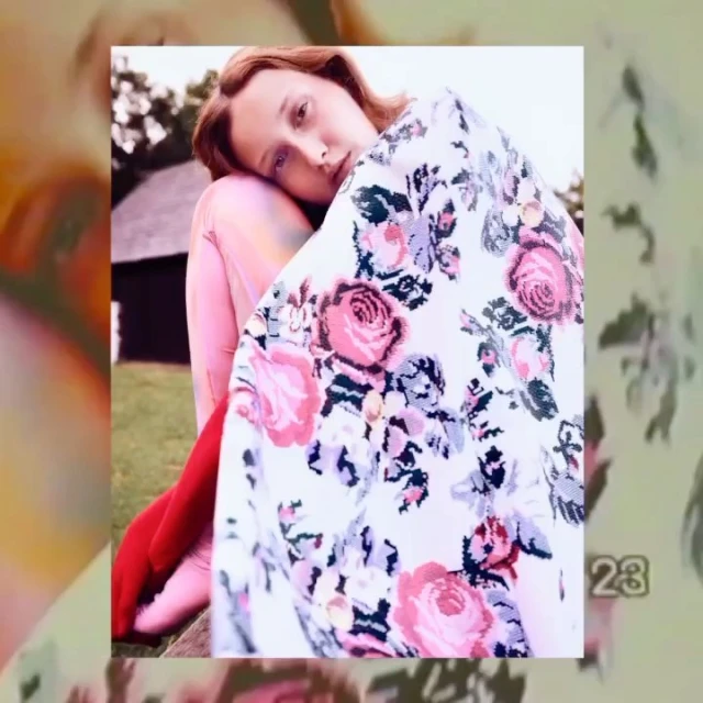 Woman sits outdoors wrapped in floral blanket. Medium shot portrait with natural light and a classic aesthetic.