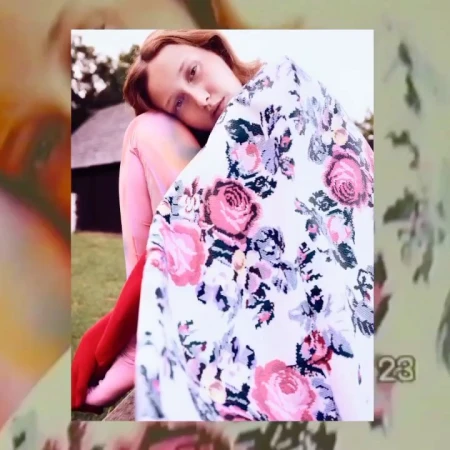 Woman sits outdoors wrapped in floral blanket. Medium shot portrait with natural light and a classic aesthetic.