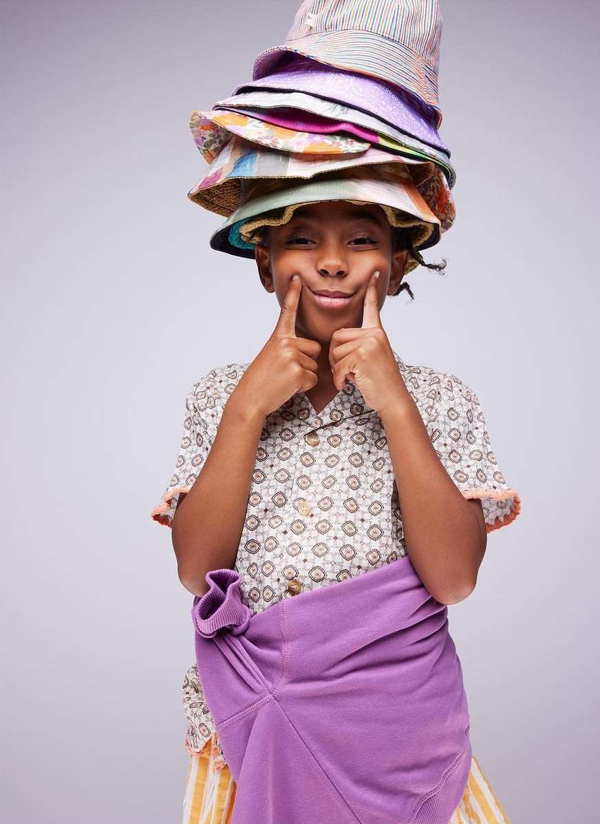 Young boy with several colorful hats stacked on his head, holding a purple sweatshirt, making a silly face in a studio shot.