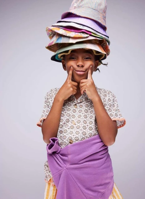 Young boy with several colorful hats stacked on his head, holding a purple sweatshirt, making a silly face in a studio shot.