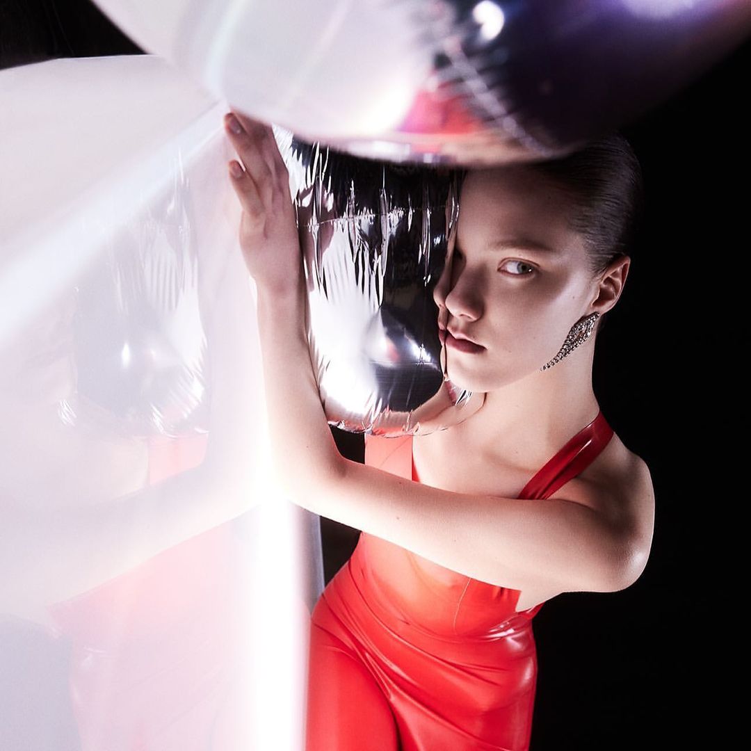 Glamorous woman in a red dress poses with silver and transparent balloons in a studio with a dark background, showcasing an earring.