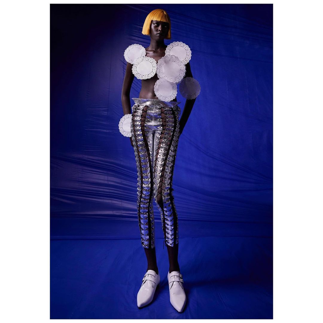 A dark-skinned model with a yellow bob stands against a deep blue backdrop, wearing paper doilies as a top and metallic leggings.