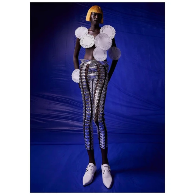 A dark-skinned model with a yellow bob stands against a deep blue backdrop, wearing paper doilies as a top and metallic leggings.