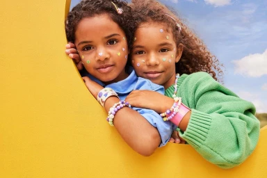 Two children with colorful face stickers and beaded bracelets leaning against a bright yellow structure outdoors.