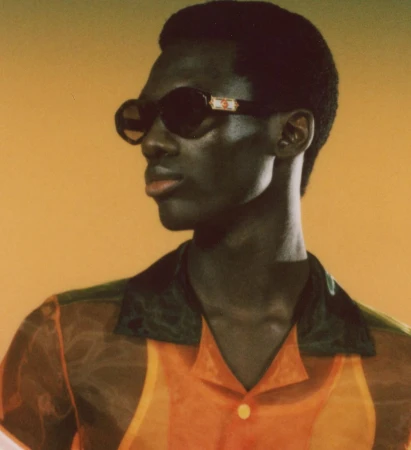 Portrait of a young Black man wearing sunglasses and a sheer orange shirt, posing against a mustard-yellow background in studio lighting.