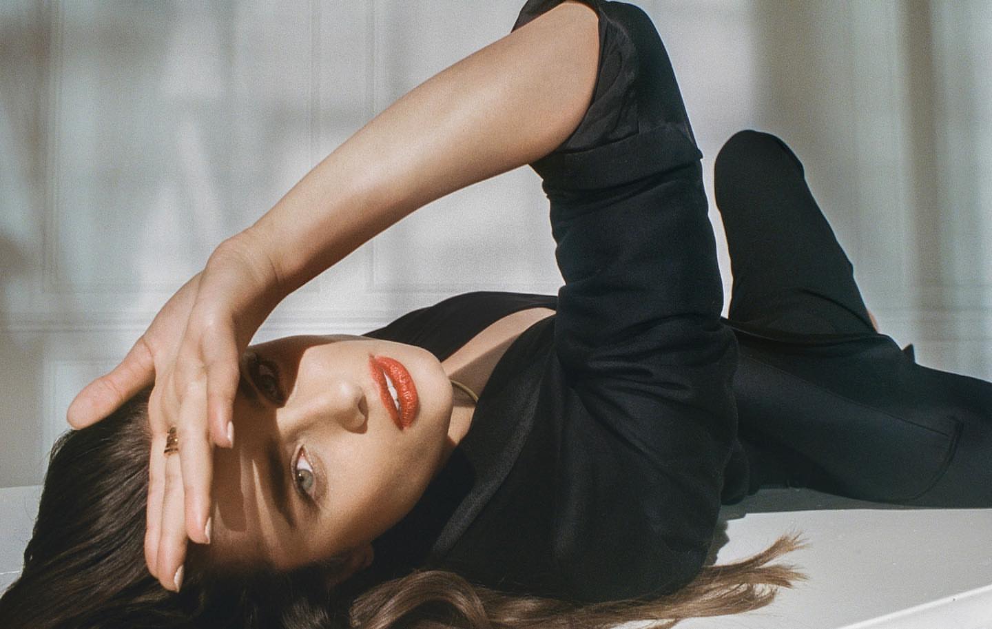 Woman in dark blazer reclining against a pale paneled wall with bright sunlight casting shadows.