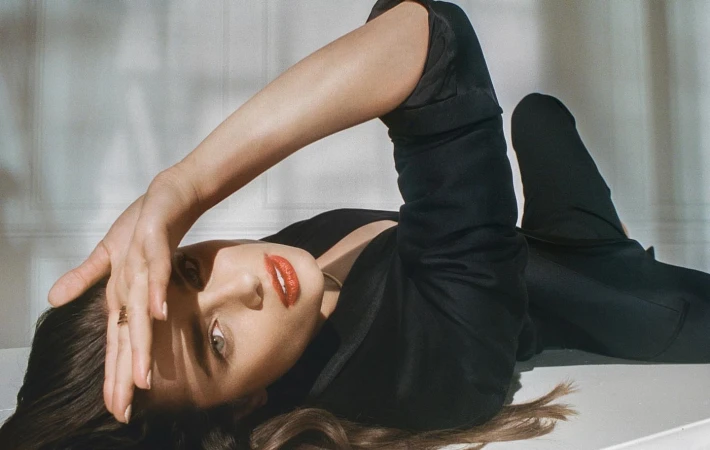 Woman in dark blazer reclining against a pale paneled wall with bright sunlight casting shadows.