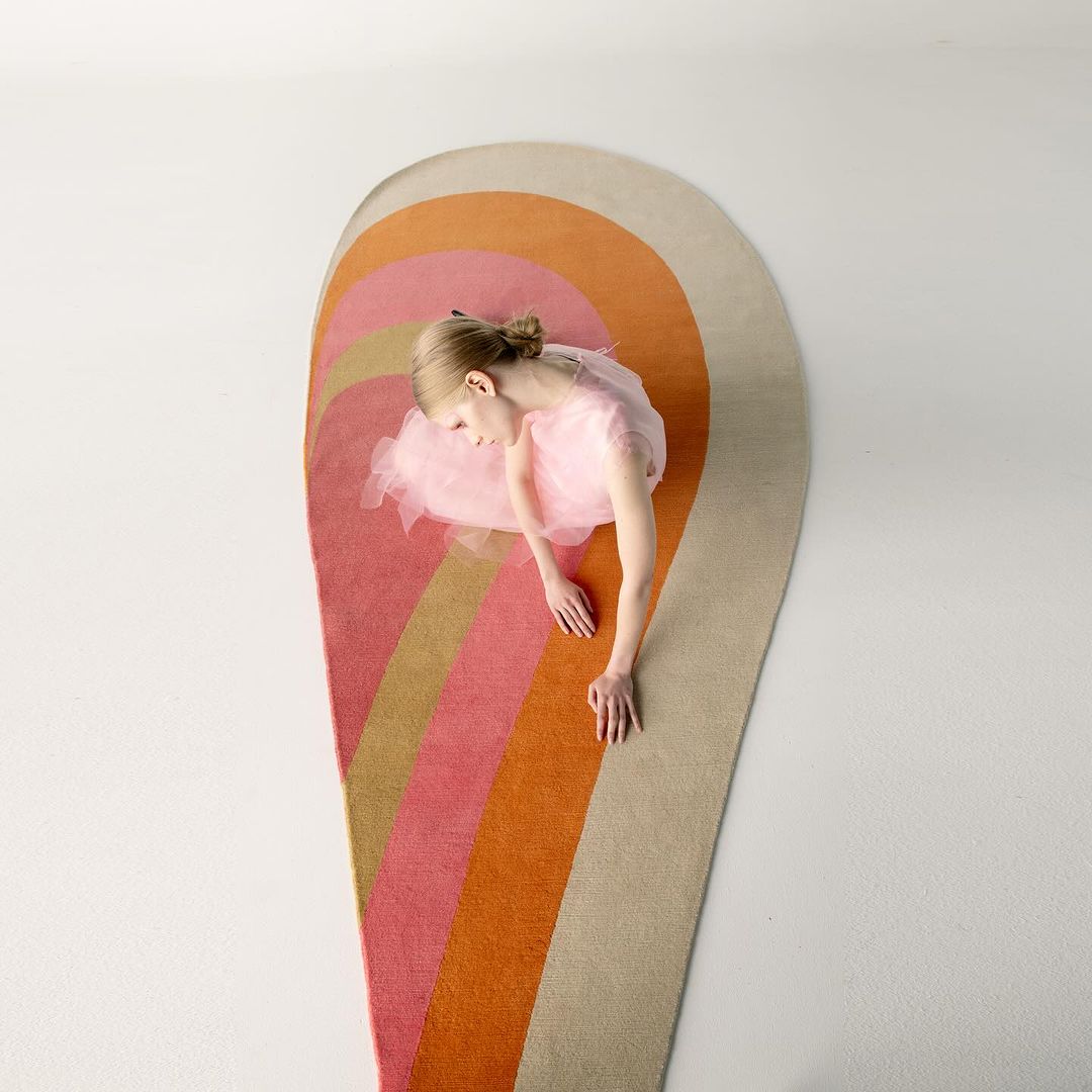 Overhead view of a young girl in a pink dress lying on a colourful striped rug against a white background, studio lit.