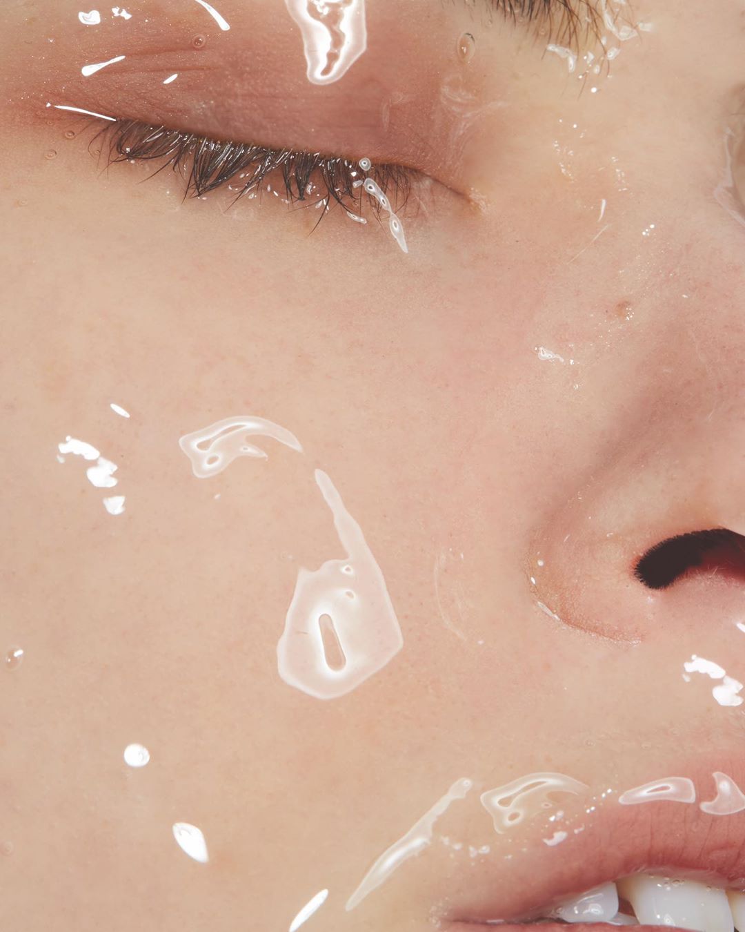 Close-up portrait of a fair-skinned woman with water droplets on her face, eyes closed, showcasing beauty and skincare in soft lighting.