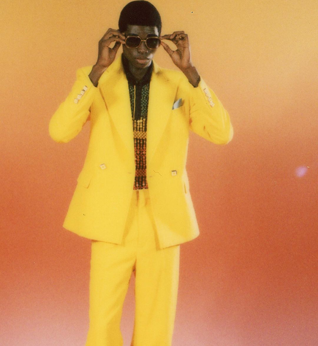An adult Black male in a bright yellow suit poses while adjusting dark sunglasses against an orange gradient backdrop in a fashion shot.