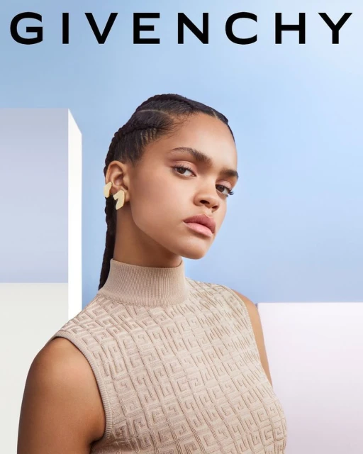 Close-up of a woman with braided hair wearing gold earrings and a beige patterned top against a pale blue and white backdrop.