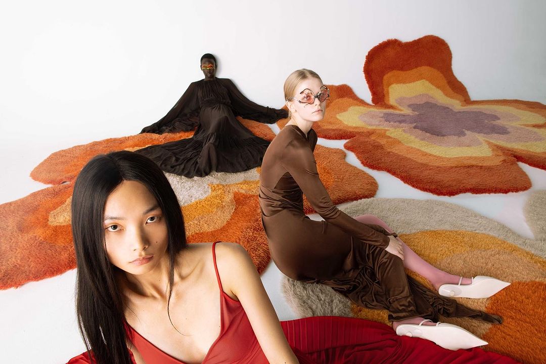 Three models wearing high fashion dresses posed on large colorful organic-shaped rugs against a white studio background.
