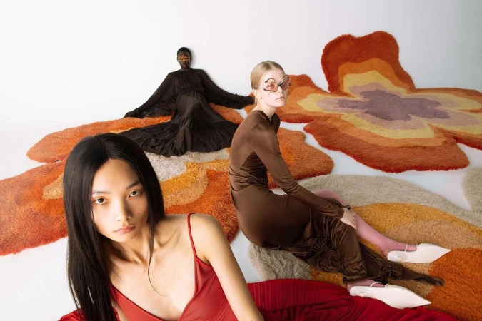 Three models wearing high fashion dresses posed on large colorful organic-shaped rugs against a white studio background.