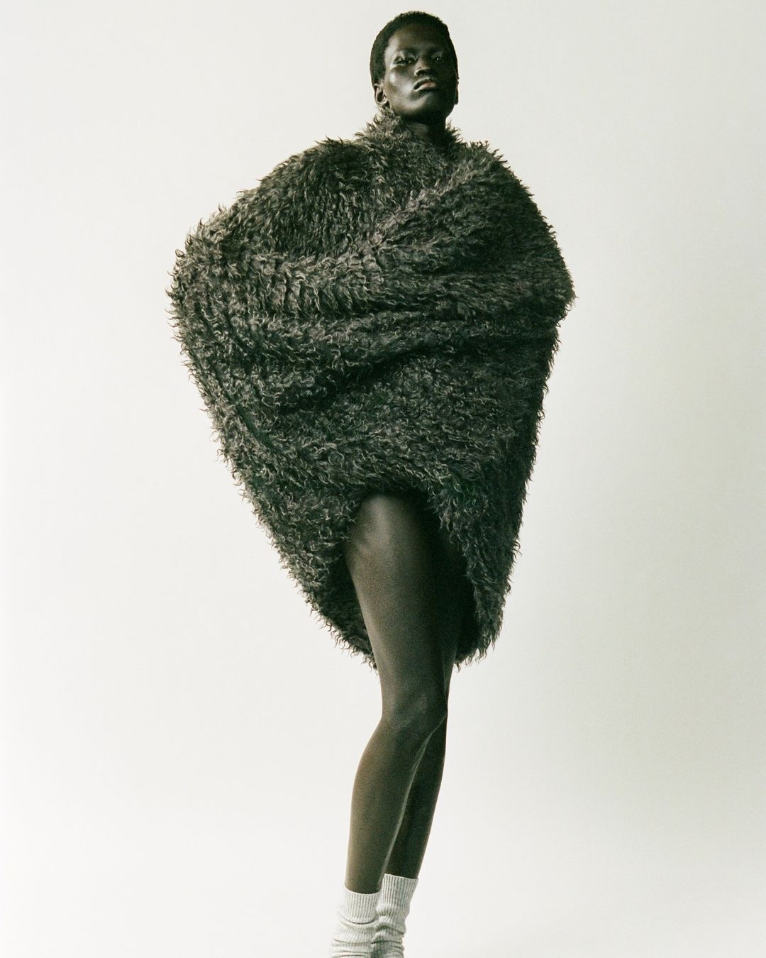 Fashion shot of a slender, dark-skinned woman standing in a photo studio, wearing a furry oversized jacket and knit socks.