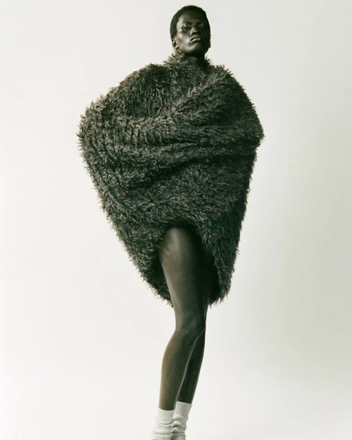 Fashion shot of a slender, dark-skinned woman standing in a photo studio, wearing a furry oversized jacket and knit socks.