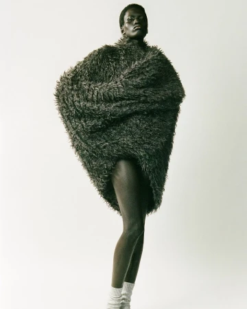 Fashion shot of a slender, dark-skinned woman standing in a photo studio, wearing a furry oversized jacket and knit socks.