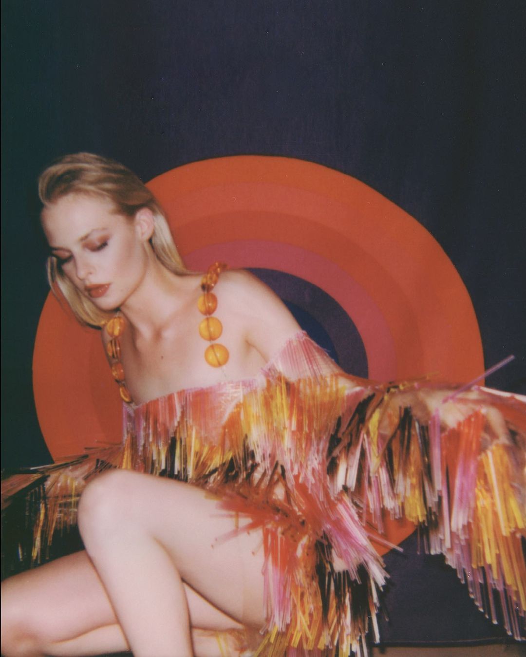 Woman poses in a fringed dress against a dark backdrop with an orange, red, and brown concentric circle design, fashion photography.