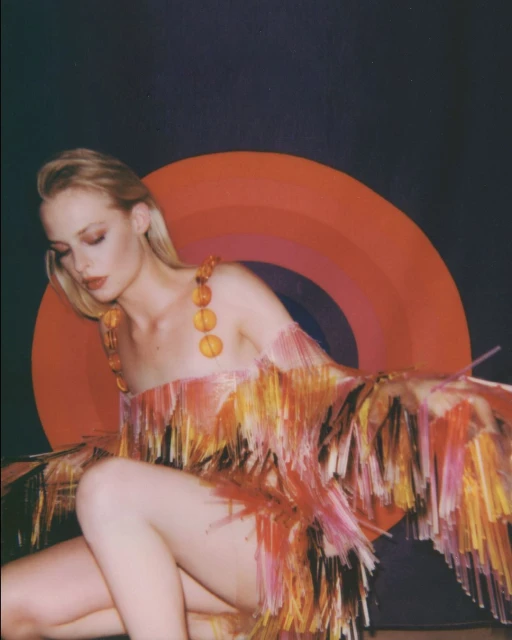 Woman poses in a fringed dress against a dark backdrop with an orange, red, and brown concentric circle design, fashion photography.