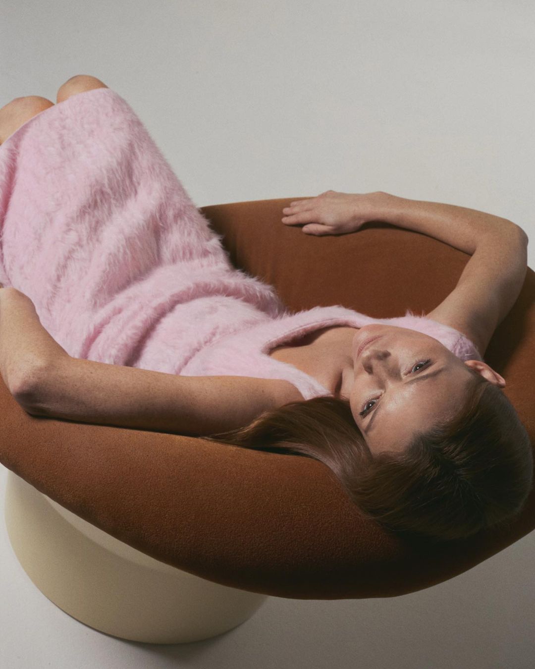 Woman in pink dress lounges in a tan chair against a white backdrop, showcasing fashion and design elements in a studio setting.