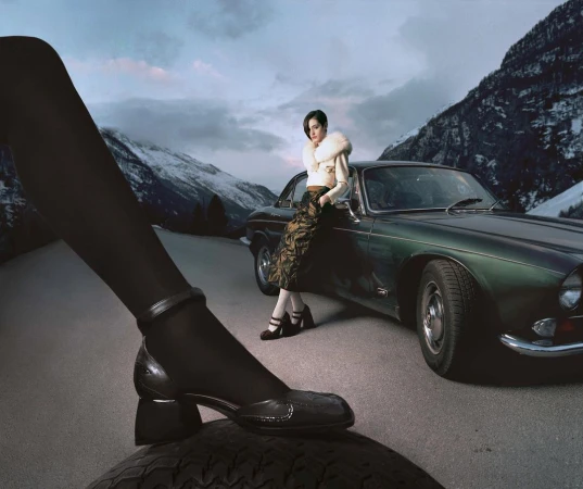 Model leaning against a dark green vintage car with snowy mountain peaks in the background by photographer Damien Krisl