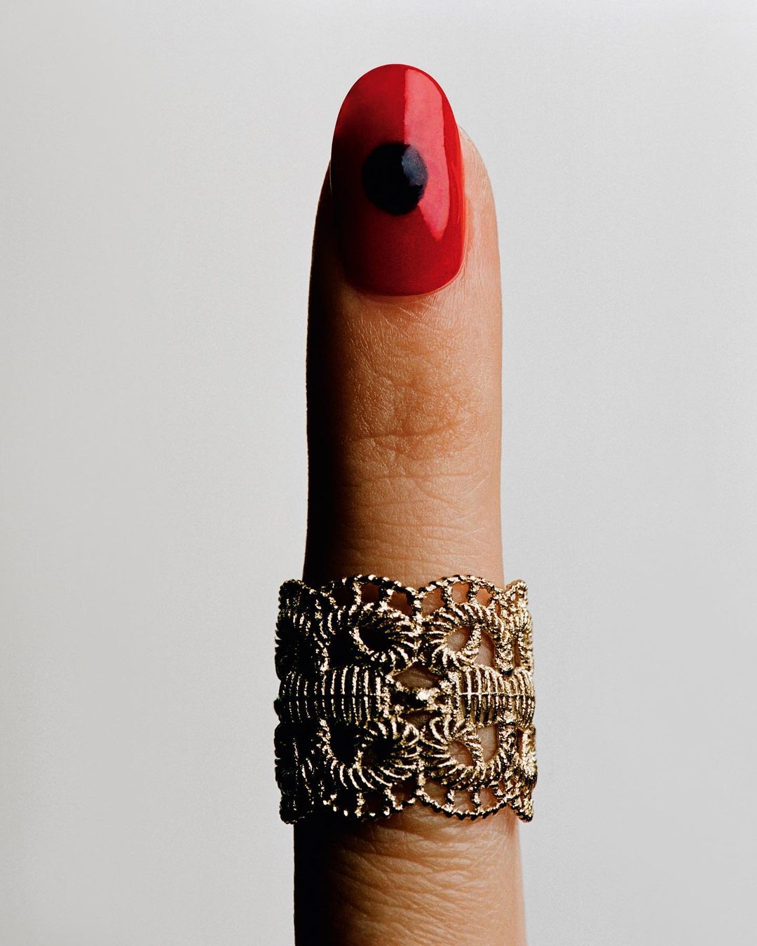 Close-up of a finger with a gold metal filigree ring and a red fingernail with a black circle design against a neutral background.