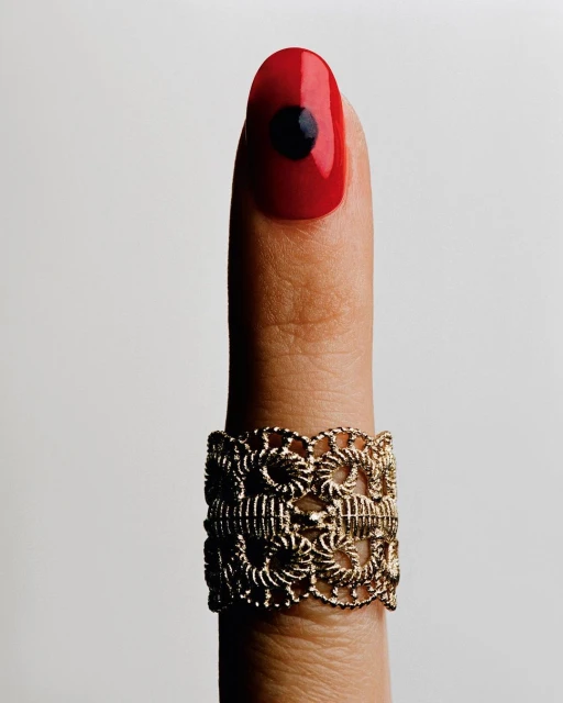 Close-up of a finger with a gold metal filigree ring and a red fingernail with a black circle design against a neutral background.
