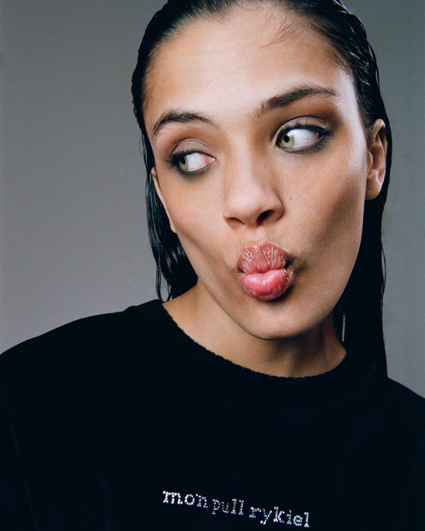 Close-up portrait of a young woman with wet hair, looking off to the side with puckered lips, wearing a black sweater with text.