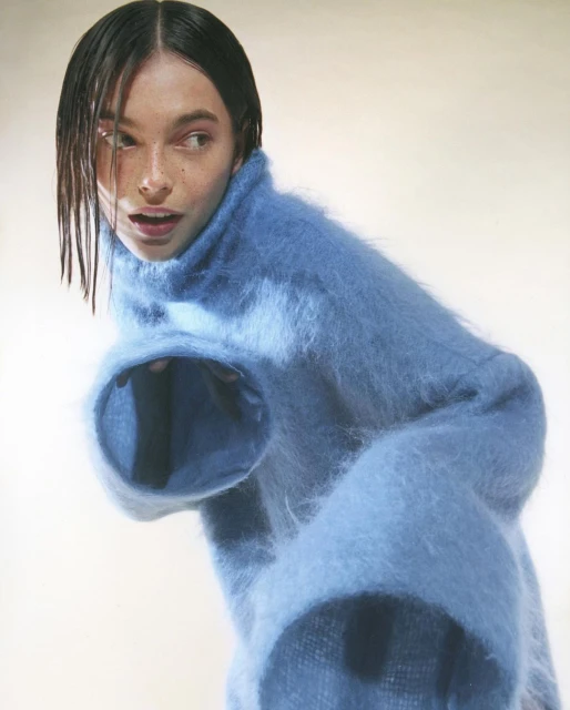 A fair-skinned young woman poses in a baby-blue fuzzy sweater, looking to the left with a neutral expression in a photo studio.