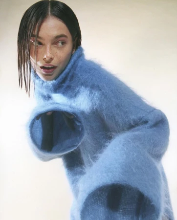 A fair-skinned young woman poses in a baby-blue fuzzy sweater, looking to the left with a neutral expression in a photo studio.