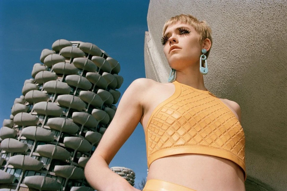 Woman in yellow halter top and oversized earrings poses in front of the residential building with globular balcony extensions on a sunny day.