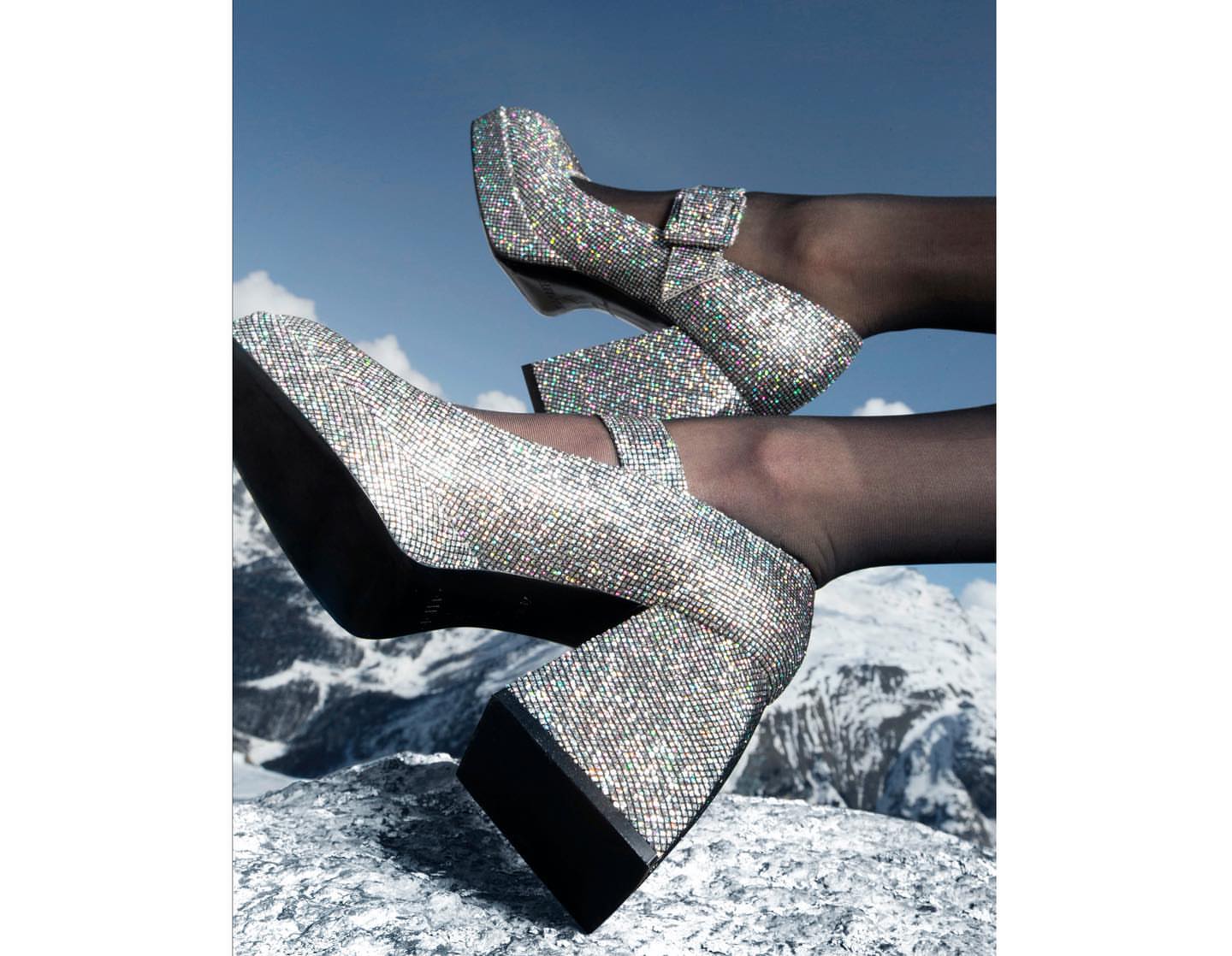Two pairs of legs wearing shimmering platform Mary Jane heels are positioned on a textured rock surface against a mountain and sky backdrop.
