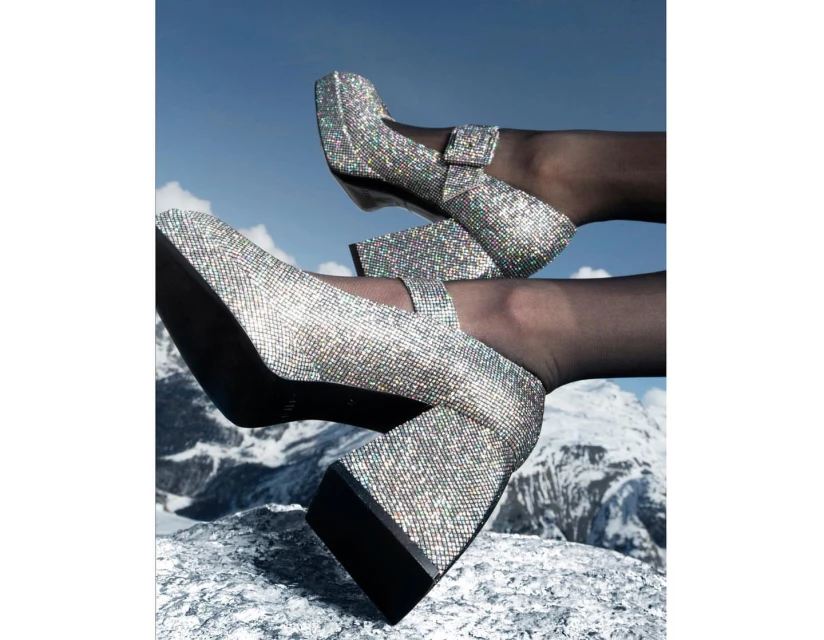 Two pairs of legs wearing shimmering platform Mary Jane heels are positioned on a textured rock surface against a mountain and sky backdrop.